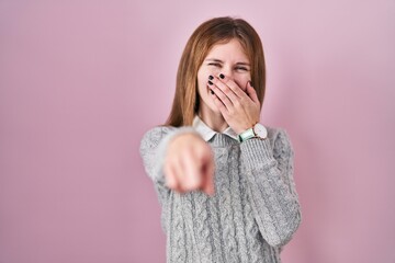 Beautiful woman standing over pink background laughing at you, pointing finger to the camera with hand over mouth, shame expression