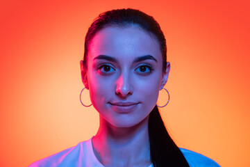 Closeup face of young attractive girl looking at camera isolated on orange color background in neon light. Concept of emotions, facial expressions
