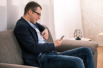 Portrait side view of caucasian man wearing jacket and glasses sitting on sofa in open position looking to phone against of alarge window with curtain. Success and business concept