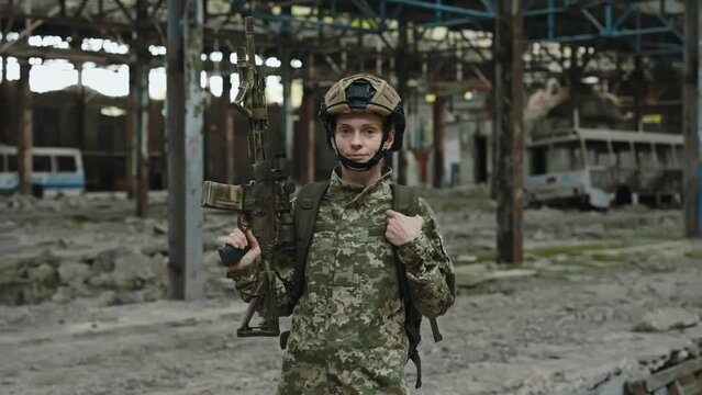 Portrait of fearless female fighter with weapon in hands confidently looking at camera while standing on industrial plant. Professionally trained soldier patrolling at destroyed factory during war.