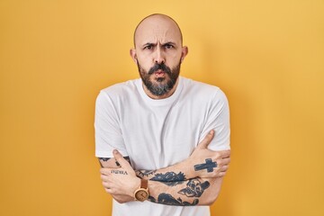 Young hispanic man with tattoos standing over yellow background shaking and freezing for winter...