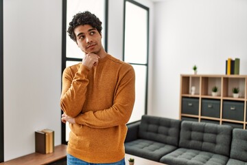 Young hispanic man wearing casual clothes standing at home serious face thinking about question with hand on chin, thoughtful about confusing idea