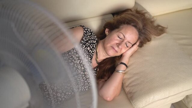 Woman lying on a sofa and giving fresh air from a fan because of the high temperatures, slowmotion shot.