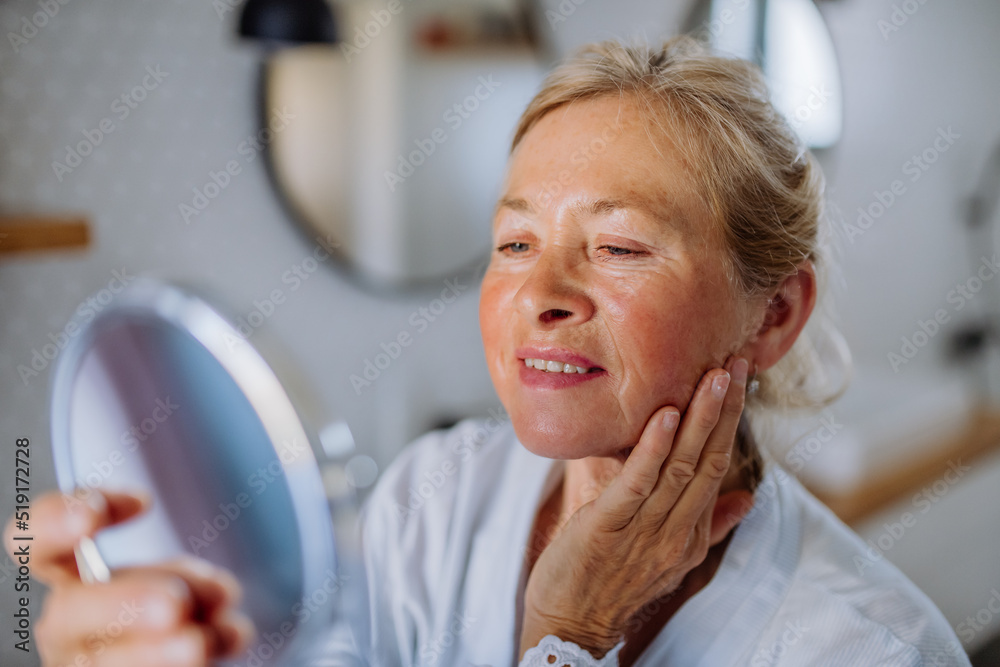 Wall mural Beautiful senior woman in bathrobe looking at mirror and applying natural face cream in bathroom, skin care concept.