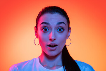 Closeup face of young attractive girl looking at camera isolated on orange color background in neon light. Concept of emotions, facial expressions