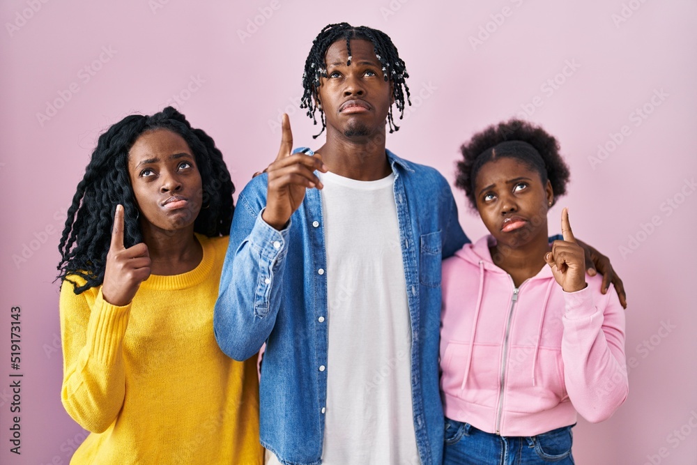 Sticker group of three young black people standing together over pink background pointing up looking sad and