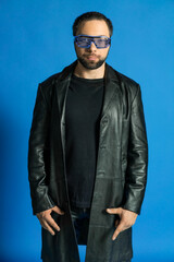 Obraz premium portrait of a man in a studio in a black leather coat. model with a beard on a blue background. trendy luminous glasses on the face
