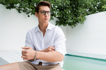 man with silver bracelet and eyeglasses sitting near pool and looking away.