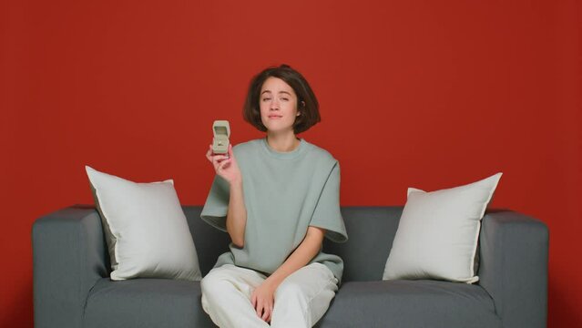 Young woman opens gift box with engagement ring unpleased with marriage proposal, grimacing, sitting on couch