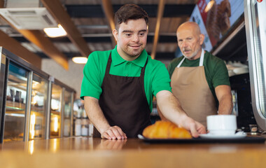 Happy waiter with Down syndrome serving coffee with help of his collegue at cafe.