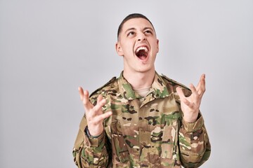 Young man wearing camouflage army uniform crazy and mad shouting and yelling with aggressive expression and arms raised. frustration concept.