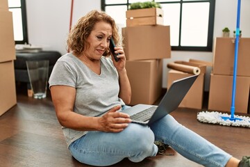 Fototapeta premium Middle age caucasian woman using laptop and talking on the smartphone sitting on the floor at new home.