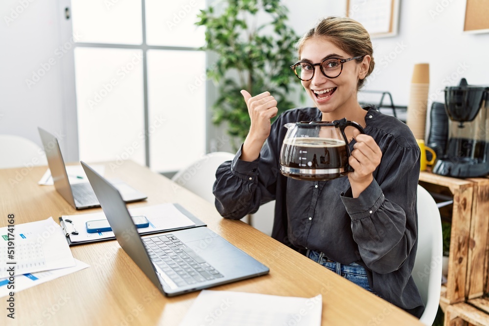 Poster young blonde woman working at the office holding coffee pot pointing thumb up to the side smiling ha