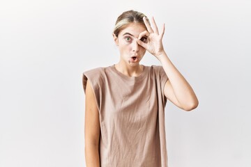 Young blonde woman standing over isolated background doing ok gesture shocked with surprised face, eye looking through fingers. unbelieving expression.