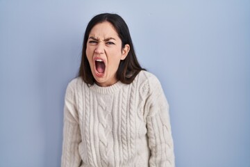 Young brunette woman standing over blue background angry and mad screaming frustrated and furious,...