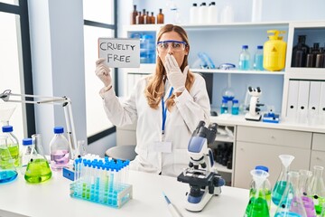 Young blonde scientist woman working on cruelty free laboratory covering mouth with hand, shocked and afraid for mistake. surprised expression