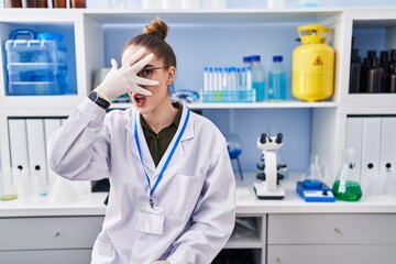 Young hispanic girl working at scientist laboratory peeking in shock covering face and eyes with...