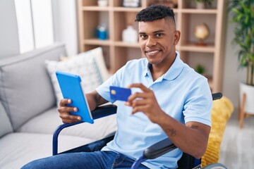 Young latin man using touchpad and credit card sitting on wheelchair at home
