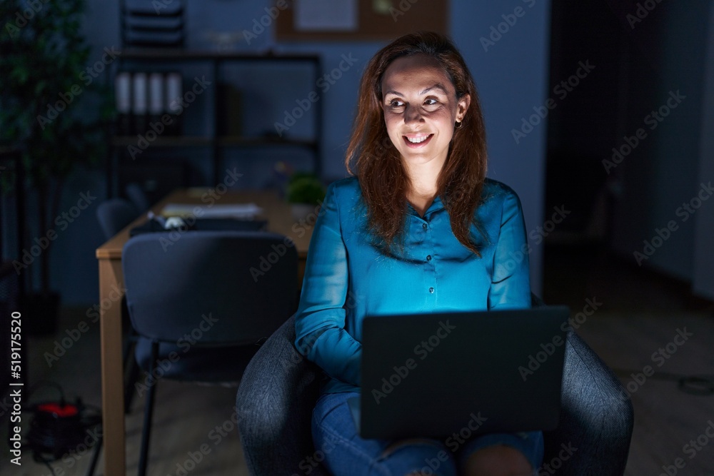 Poster brunette woman working at the office at night looking away to side with smile on face, natural expre