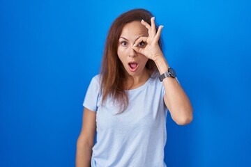 Brunette woman standing over blue background doing ok gesture shocked with surprised face, eye looking through fingers. unbelieving expression.