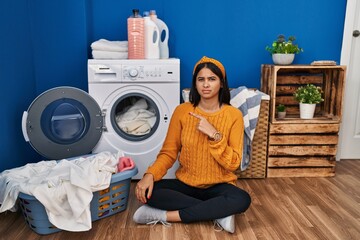 Young hispanic woman doing laundry pointing aside worried and nervous with forefinger, concerned...