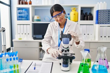Young latin woman wearing scientist uniform using microscope at laboratory