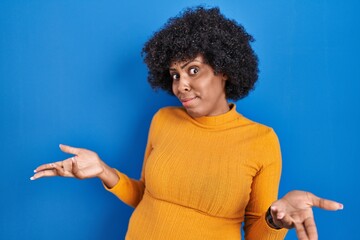 Black woman with curly hair standing over blue background clueless and confused with open arms, no idea concept.