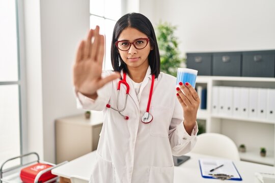 Young hispanic doctor woman holding cotton buds with open hand doing stop sign with serious and confident expression, defense gesture