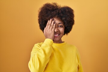 Fototapeta na wymiar Young african american woman standing over yellow background covering one eye with hand, confident smile on face and surprise emotion.