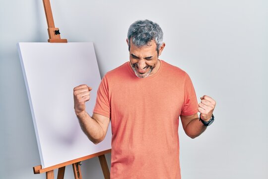 Handsome middle age man with grey hair standing by painter easel stand very happy and excited doing winner gesture with arms raised, smiling and screaming for success. celebration concept.