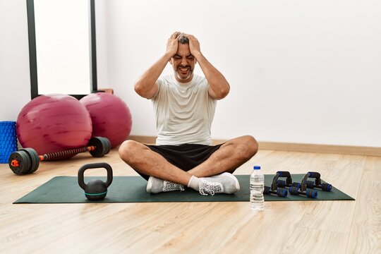 Middle Age Hispanic Man Sitting On Training Mat At The Gym Suffering From Headache Desperate And Stressed Because Pain And Migraine. Hands On Head.