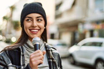 Young hispanic woman smiling happy using microphone at the city.
