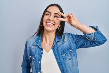 Hispanic woman standing over blue background doing peace symbol with fingers over face, smiling cheerful showing victory