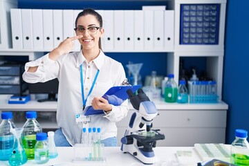 Young brunette woman working at scientist laboratory pointing with hand finger to face and nose, smiling cheerful. beauty concept