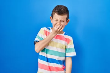 Foto op Canvas Young caucasian kid standing over blue background smelling something stinky and disgusting, intolerable smell, holding breath with fingers on nose. bad smell © Krakenimages.com