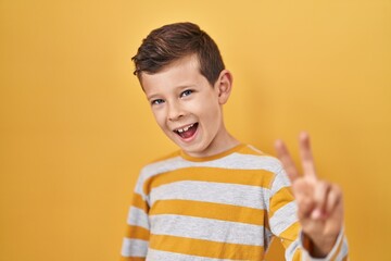 Young caucasian kid standing over yellow background smiling looking to the camera showing fingers doing victory sign. number two.