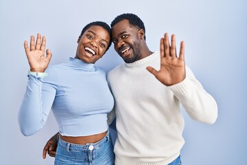 Young african american couple standing over blue background waiving saying hello happy and smiling,...
