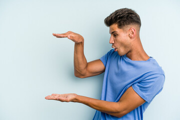 Young caucasian man isolated on blue background shocked and amazed holding a copy space between hands.