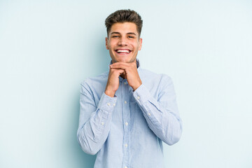 Young caucasian man isolated on blue background keeps hands under chin, is looking happily aside.