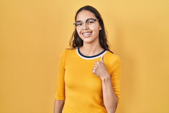 Young brazilian woman wearing glasses over yellow background smiling happy and positive, thumb up doing excellent and approval sign
