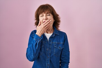 Middle age woman standing over pink background bored yawning tired covering mouth with hand. restless and sleepiness.