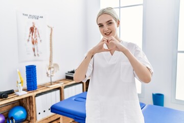 Young caucasian woman working at pain recovery clinic smiling in love doing heart symbol shape with hands. romantic concept.