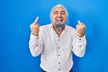 Middle age man with grey hair standing over blue background showing middle finger doing fuck you...
