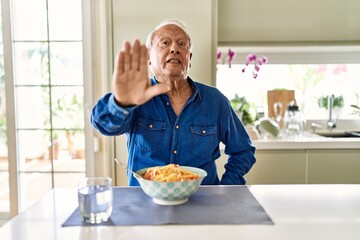 Senior man with grey hair eating pasta spaghetti at home doing stop sing with palm of the hand....