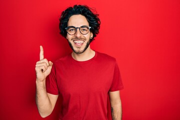 Handsome hispanic man wearing casual t shirt and glasses showing and pointing up with finger number one while smiling confident and happy.