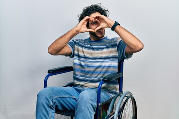 Handsome hispanic man sitting on wheelchair doing heart shape with hand and fingers smiling looking...