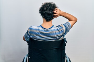 Handsome hispanic man sitting on wheelchair backwards thinking about doubt with hand on head
