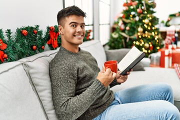 Young hispanic man reading book drinking coffee sitting on sofa by christmas tree at home