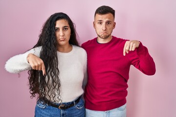 Young hispanic couple standing over pink background pointing down looking sad and upset, indicating direction with fingers, unhappy and depressed.