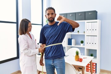 Dietitian at the clinic using tape measure measuring waist of client with angry face, negative sign...